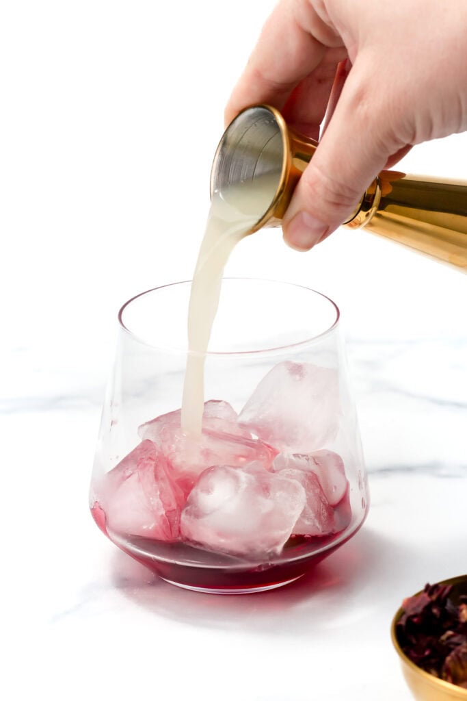A hand pours a light-colored liquid from a jigger into a glass with ice cubes and a dark red beverage, on a white surface.