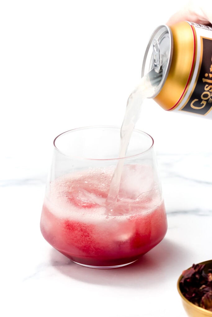 A can of soda is being poured into a glass containing a red beverage on a white surface.