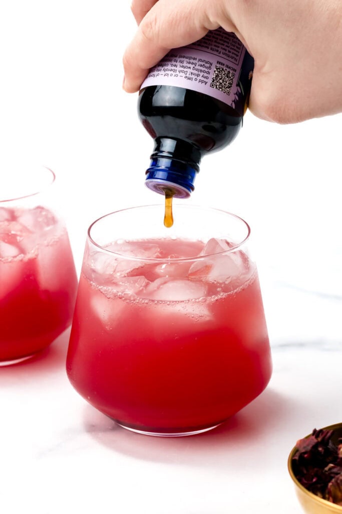 A hand pours liquid from a bottle into a glass of iced red beverage, with another similar glass and hibiscus petals nearby.