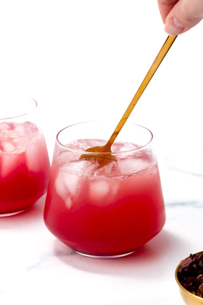 A hand stirs a pink iced drink with a gold spoon in a clear glass; another similar glass and a small bowl are nearby on a white surface.