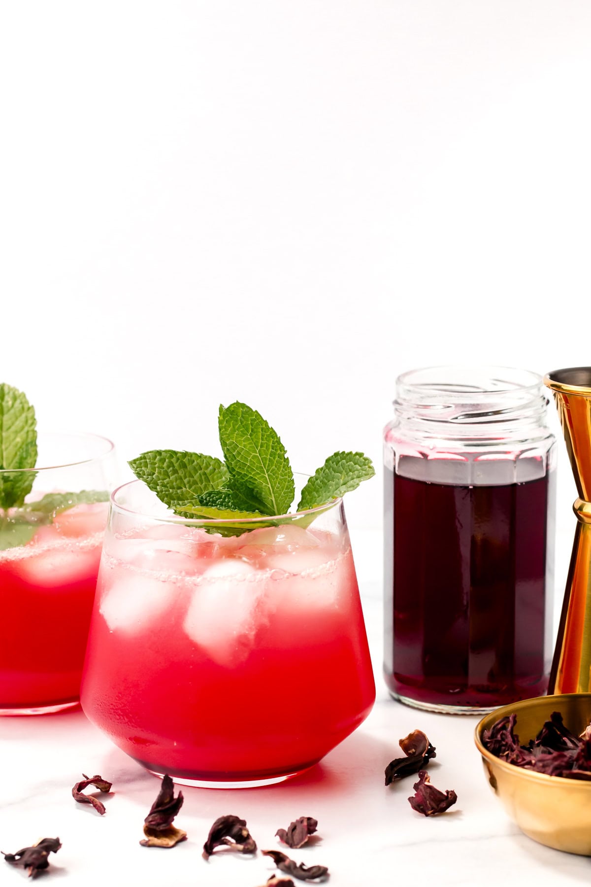 Two glasses of red iced drink garnished with mint leaves sit beside a jar filled with a dark red liquid, a gold jigger, and scattered dried hibiscus petals on a white surface.