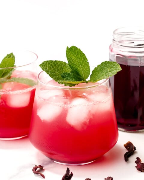 A glass of pink iced drink garnished with mint leaves, with another glass, a jar of dark liquid, and dried hibiscus petals nearby on a white surface.