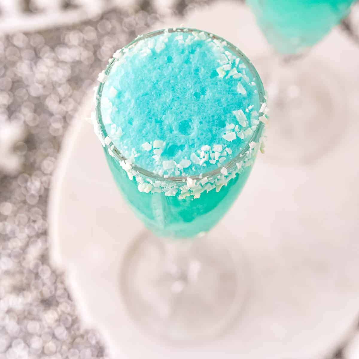 A blue frothy drink in a clear glass, with white sugar crystals on the rim, placed on a white surface.