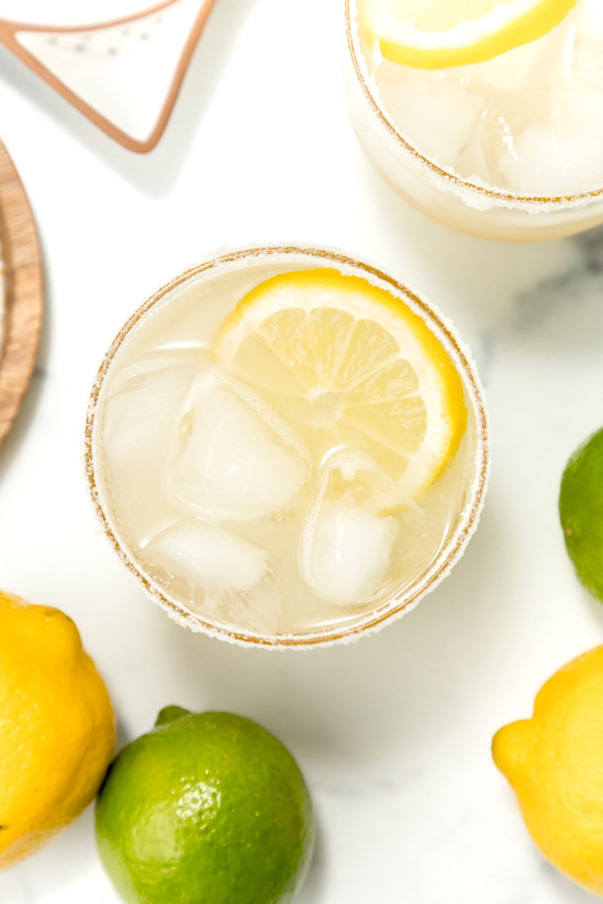 A glass of iced lemonade with a lemon slice garnish, surrounded by whole lemons and a lime on a white surface.