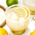 A glass of iced lemonade with a salted rim and a lemon slice, surrounded by whole lemons and limes on a white surface.