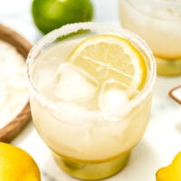 A glass of iced lemonade with a salted rim and a lemon slice, surrounded by whole lemons and limes on a white surface.
