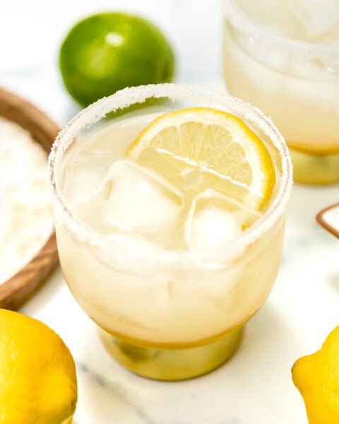 A glass of iced lemonade with a salted rim and a lemon slice, surrounded by whole lemons and limes on a white surface.