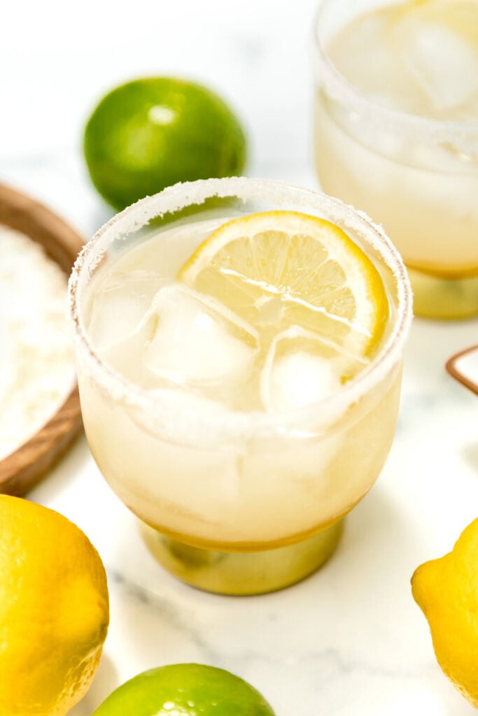 A glass of iced lemonade with a salted rim and a lemon slice, surrounded by whole lemons and limes on a white surface.