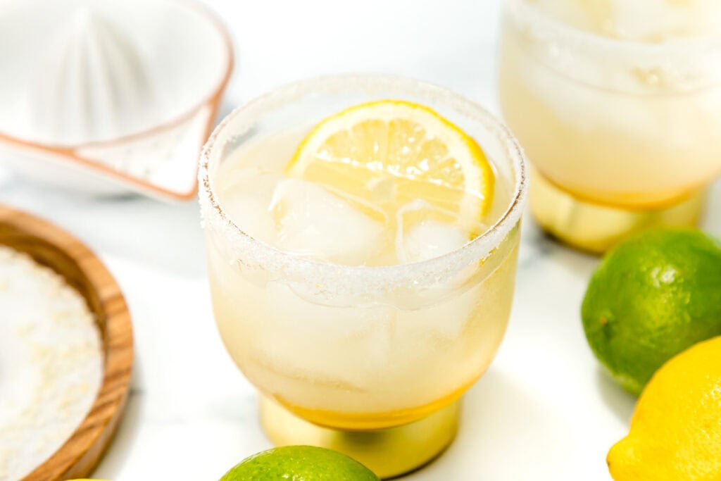 A glass of iced lemonade garnished with a lemon slice, surrounded by whole lemons and limes on a white surface.