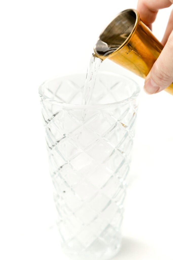 A hand pours liquid from a brass measuring jigger into a clear, diamond-patterned glass on a white background.