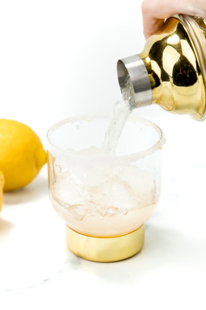 A hand pours liquid from a gold cocktail shaker into a glass with ice. Lemons are visible on the white surface nearby.