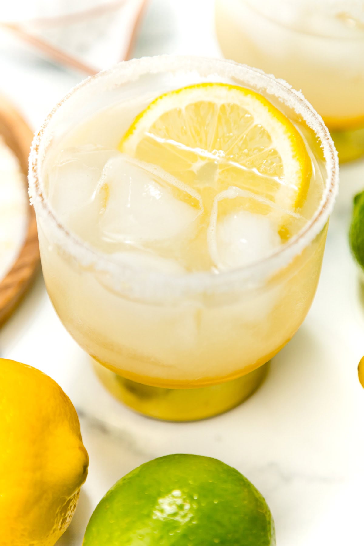 A glass with a salted rim filled with an iced lemon drink, garnished with a lemon slice; a lemon and a lime are nearby on a white surface.
