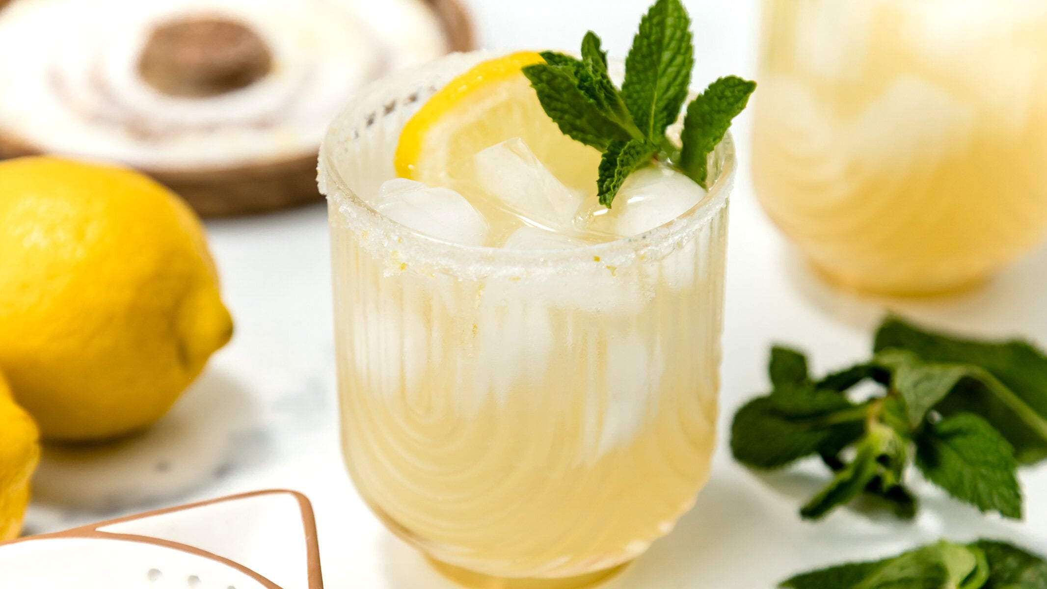 A glass of iced lemonade garnished with a lemon slice and mint leaves, with whole lemons and mint sprigs nearby.