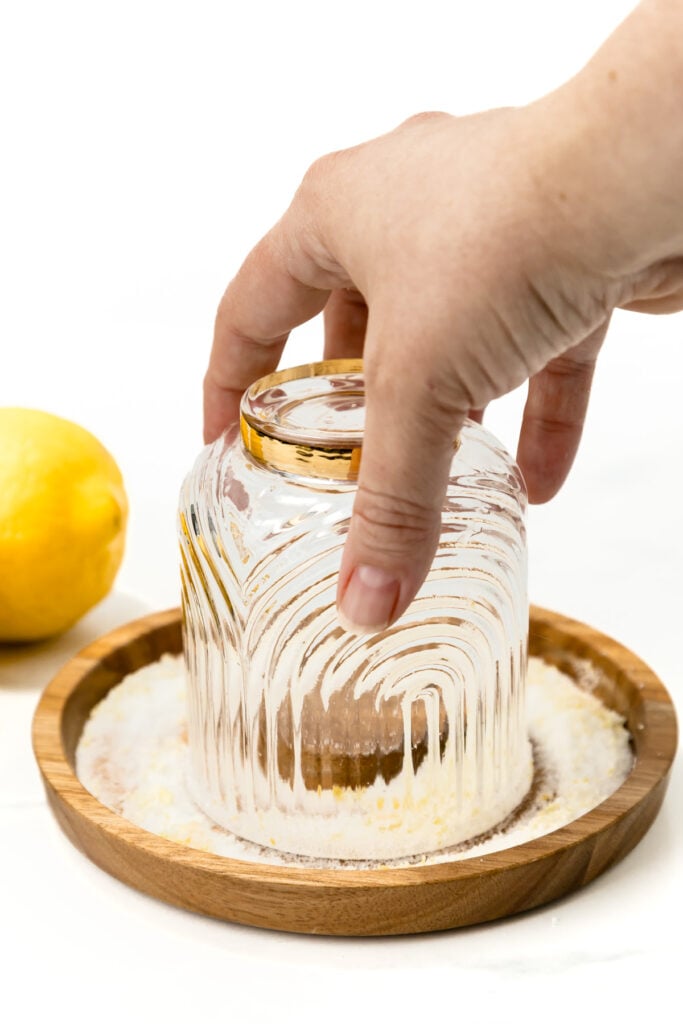 A hand presses an upside-down glass into a plate of sugar, with a lemon nearby on a white surface&mdash;perfect prep for a refreshing lemon mocktail.