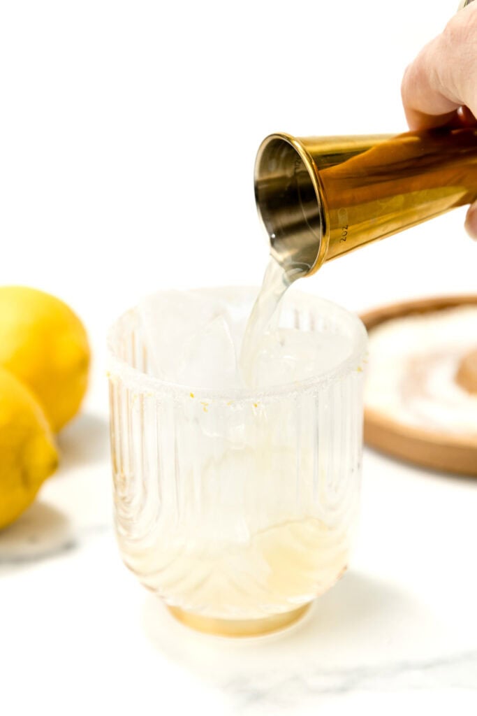 A hand pours liquid from a gold jigger into a glass with ice and a salt rim; lemons and a salt dish are in the background.