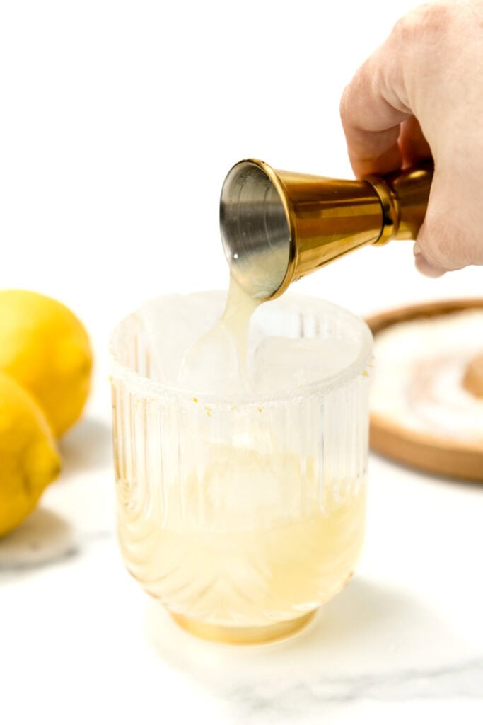 A hand pours liquid from a jigger into a glass with a salted rim, with lemons and a plate of salt in the background.