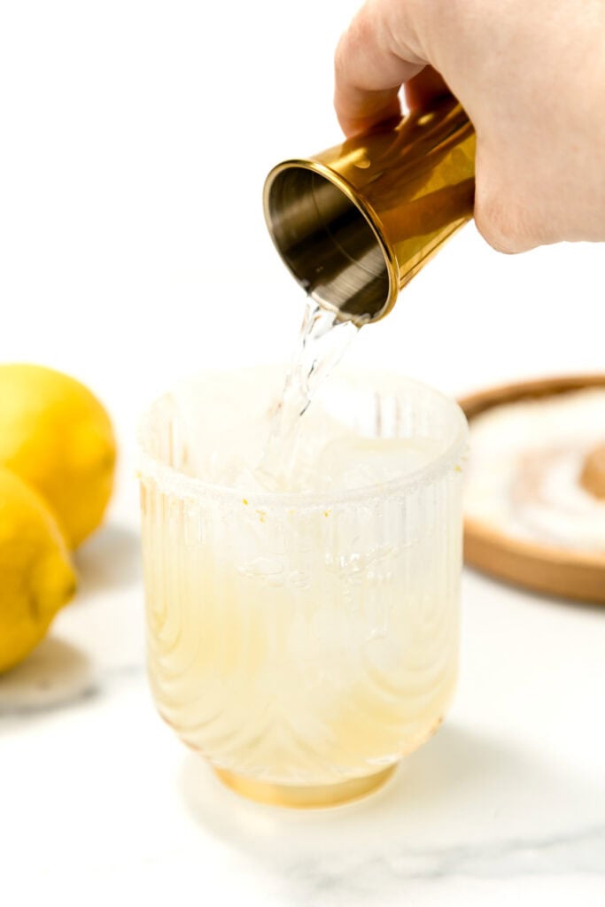 A hand pours clear liquid from a gold jigger into a glass with a salted rim; lemons and a small dish are in the background.