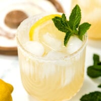 A glass of iced lemonade garnished with a lemon slice and fresh mint, with an extra lemon and mint leaves nearby on a marble surface.