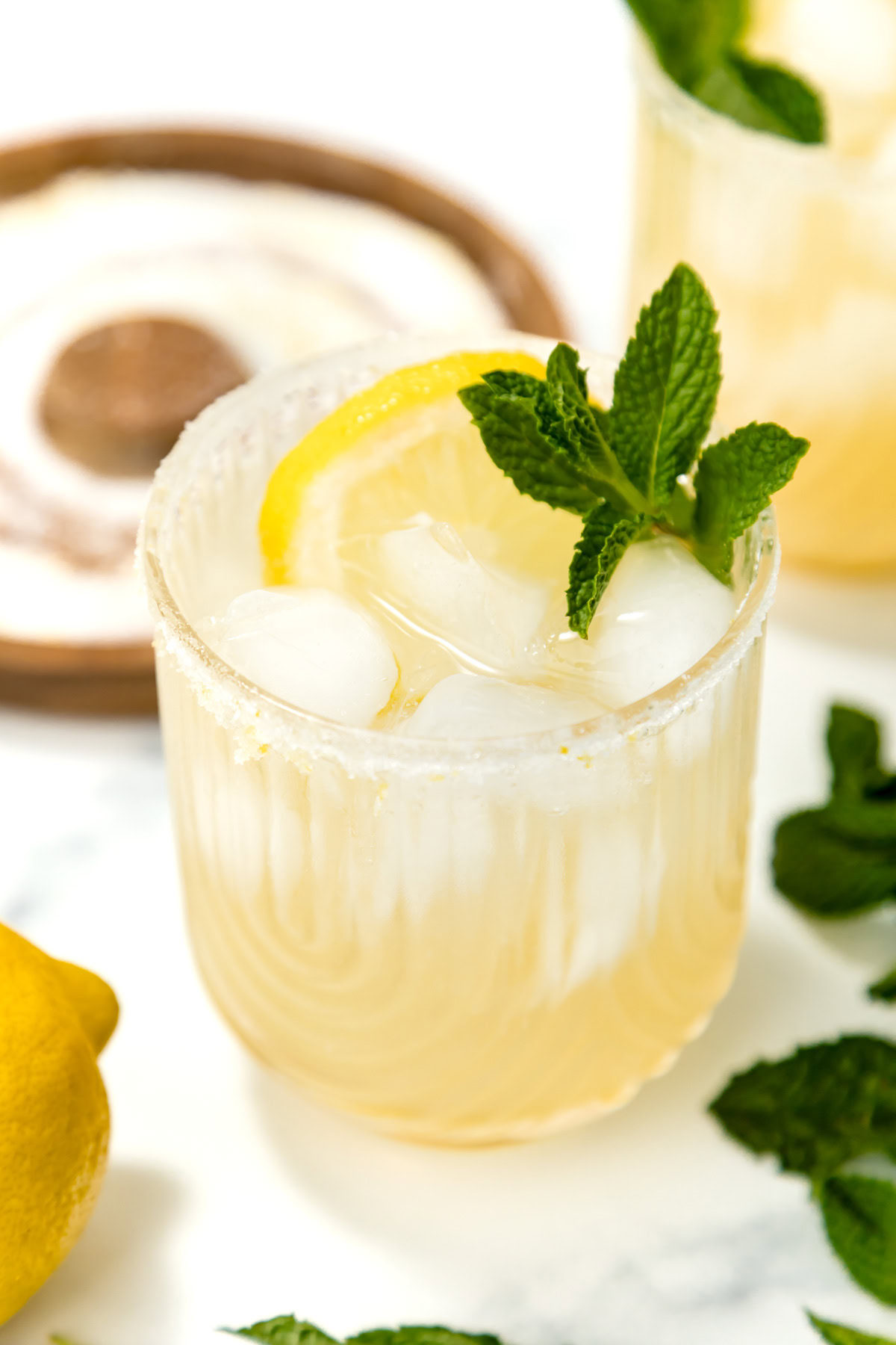 A glass of iced lemonade garnished with a lemon slice and fresh mint, with an extra lemon and mint leaves nearby on a marble surface.
