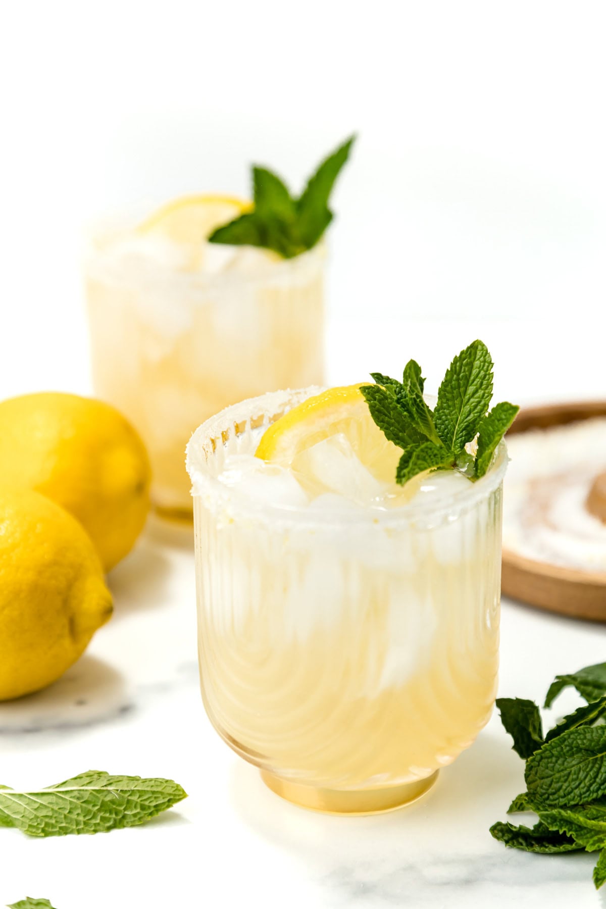 Two glasses of iced lemonade garnished with lemon slices and mint leaves, with whole lemons and mint leaves scattered nearby on a white surface.