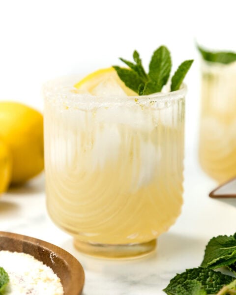 A glass of iced lemonade garnished with a lemon slice and fresh mint, with a sugared rim, sits on a white surface next to lemons and mint leaves.