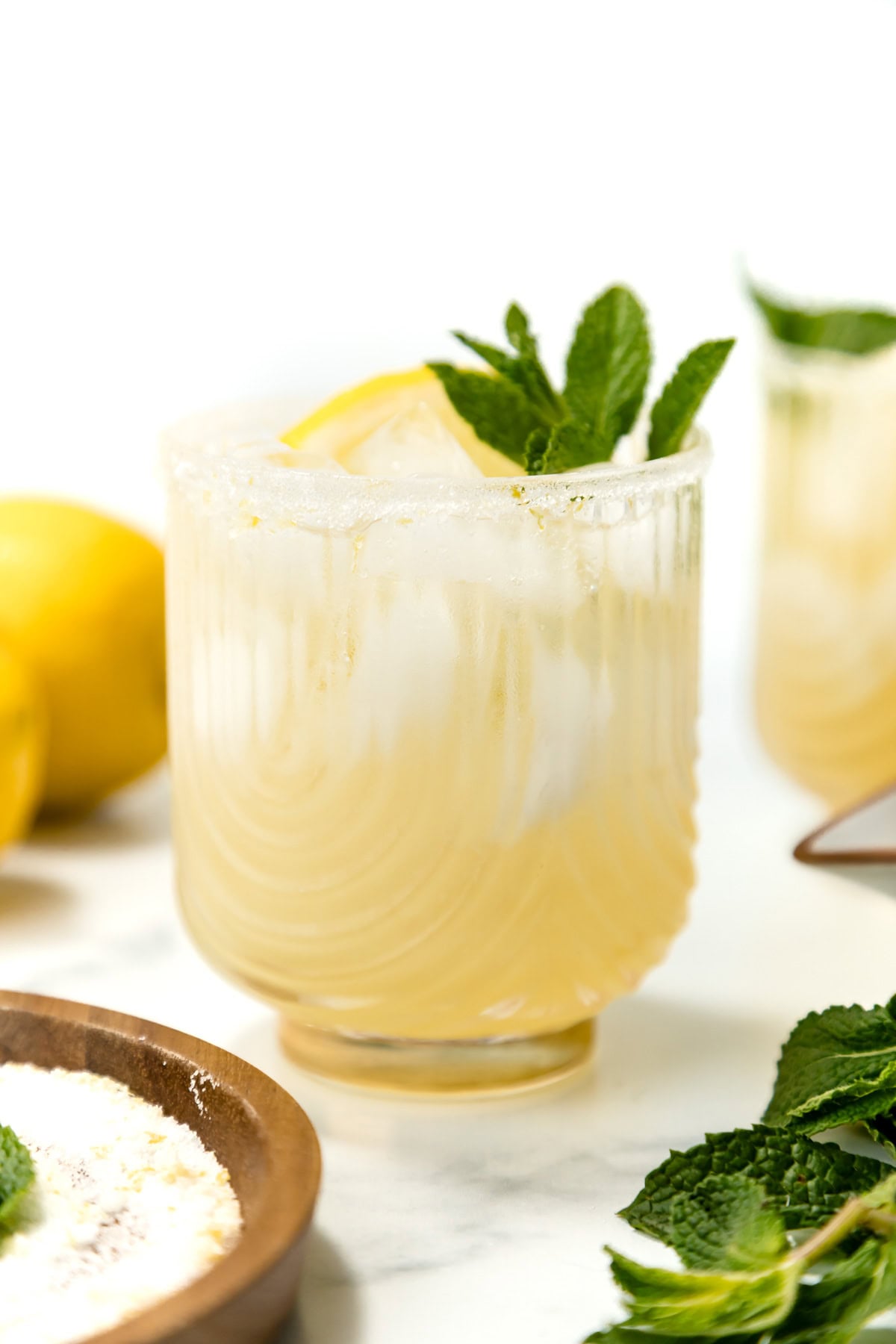 A glass of iced lemonade garnished with a lemon slice and fresh mint, with a sugared rim, sits on a white surface next to lemons and mint leaves.