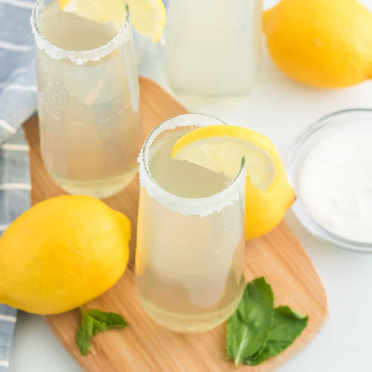 Two glasses of lemonade with lemon slices on the rim, surrounded by whole lemons, mint leaves, and a bowl of sugar on a wooden board.