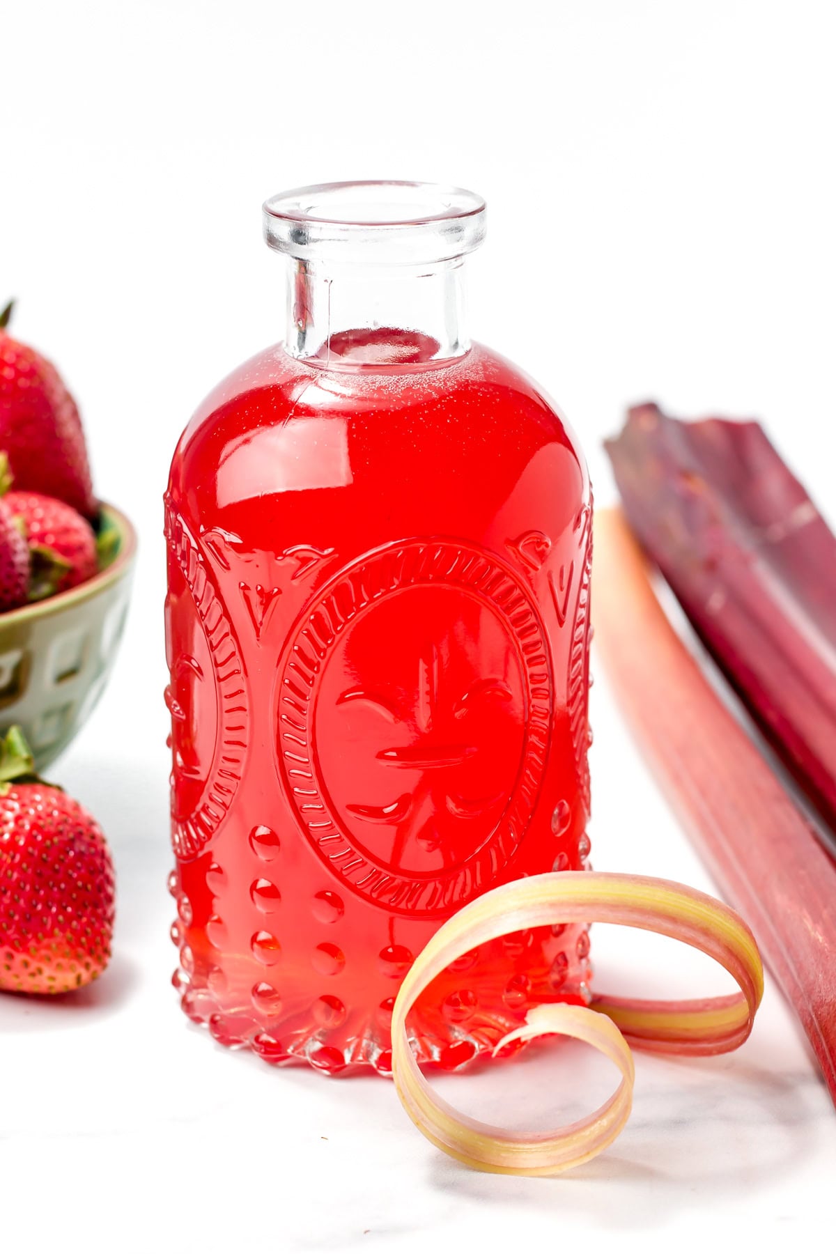 A clear glass bottle filled with bright red liquid, with strawberries, rhubarb stalks, and a curl of rhubarb placed nearby on a white surface.