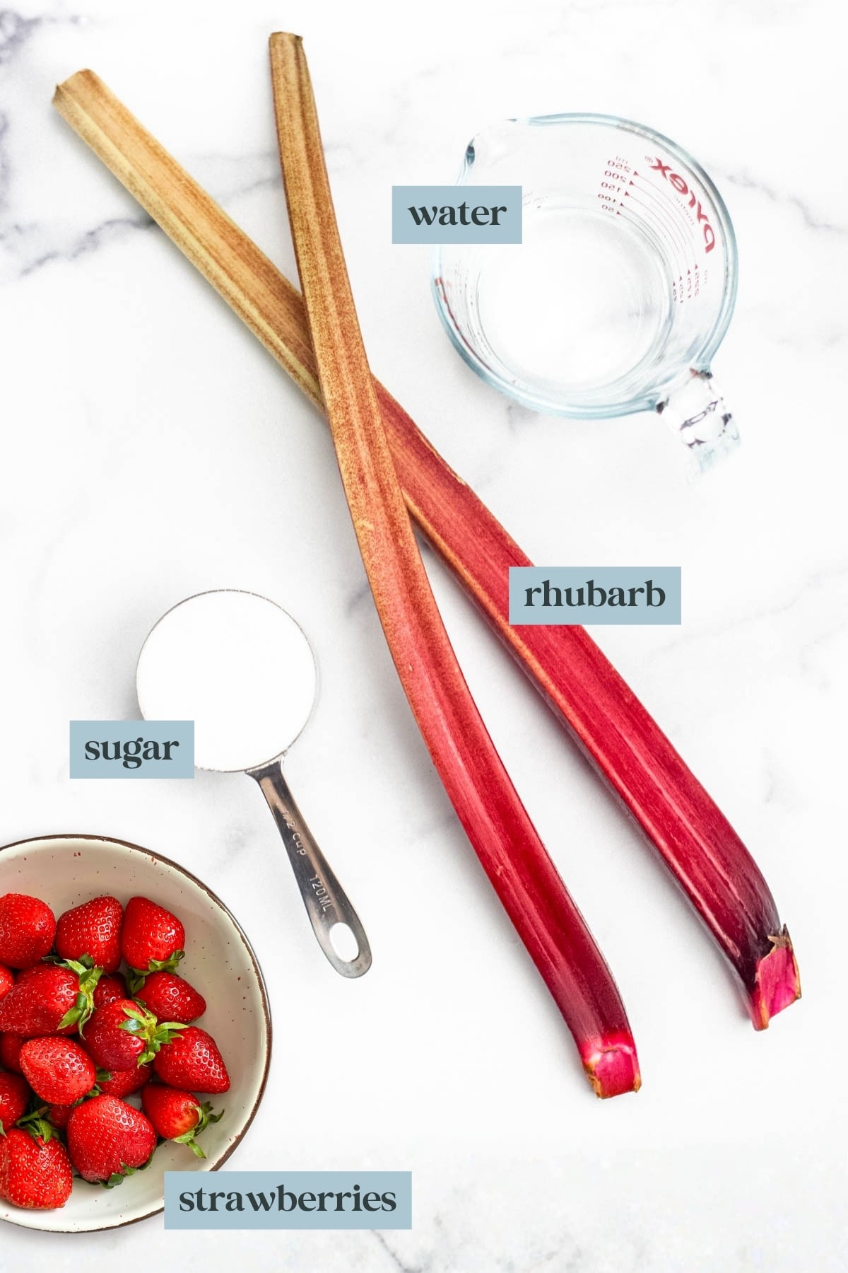 Two rhubarb stalks, a bowl of strawberries, a measuring cup of sugar, and a glass measuring cup of water on a white surface&mdash;perfect ingredients for making homemade strawberry rhubarb syrup.