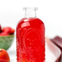 A clear glass bottle filled with bright red liquid, surrounded by strawberries, rhubarb stalks, and a green bowl of strawberries on a white surface.