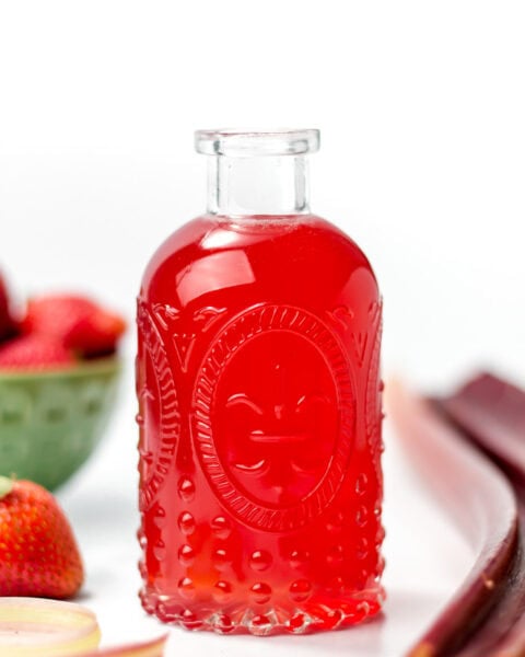 A clear glass bottle filled with bright red liquid, surrounded by strawberries, rhubarb stalks, and a green bowl of strawberries on a white surface.