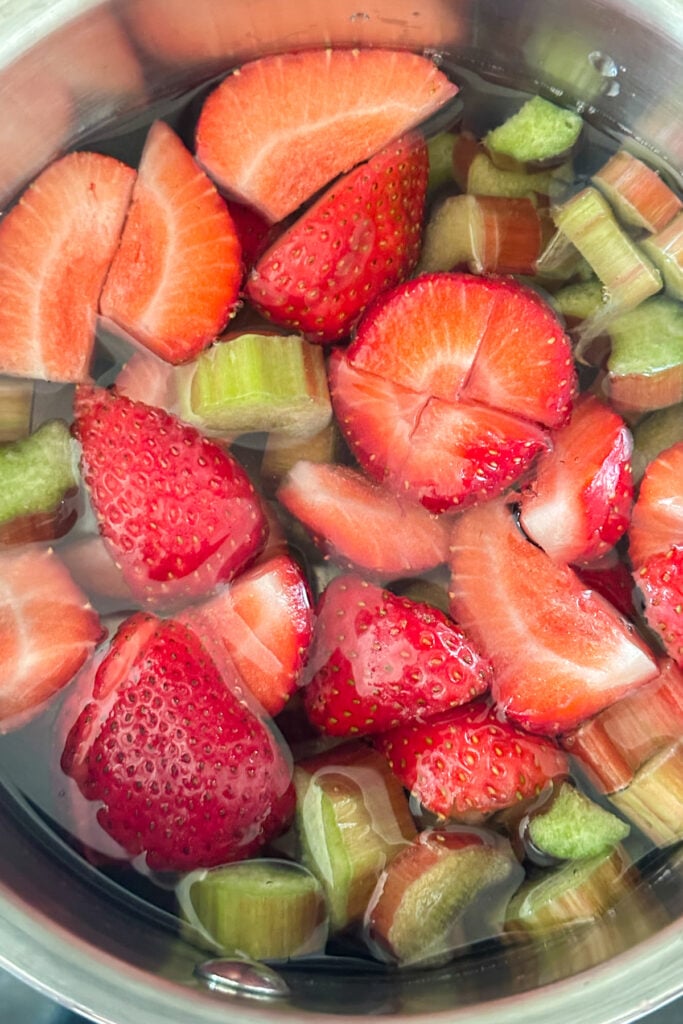 Sliced strawberries and rhubarb pieces simmer in a metal pot filled with water, ready to be transformed into a delicious strawberry rhubarb syrup.