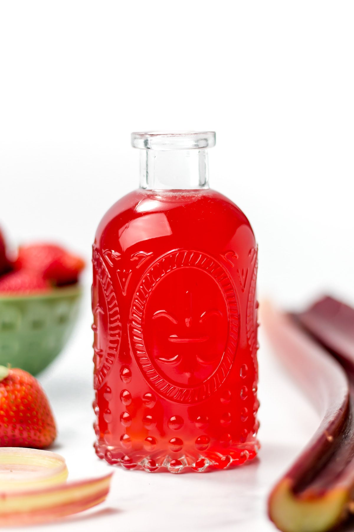 A clear glass bottle filled with bright red liquid, surrounded by strawberries, rhubarb stalks, and a green bowl of strawberries on a white surface.