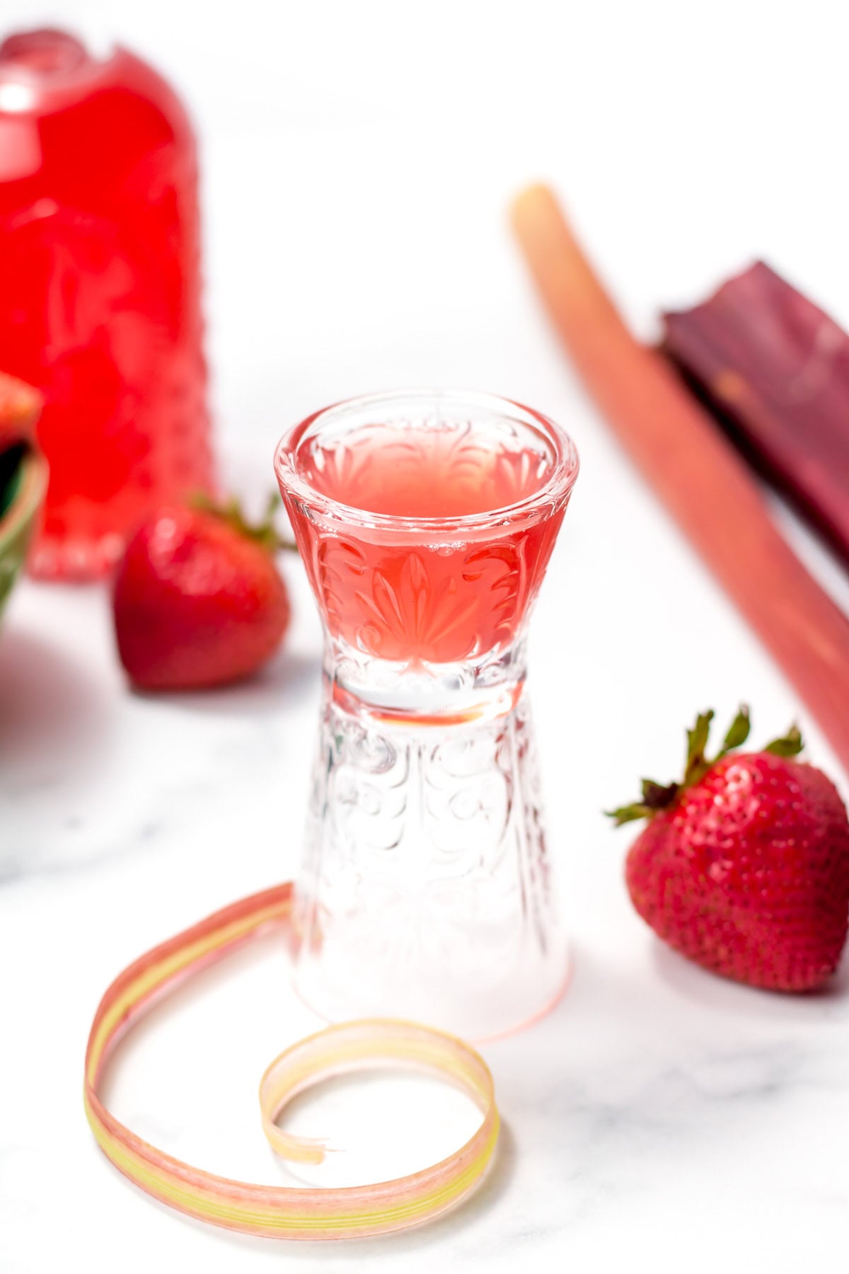A clear glass of pink liquid surrounded by strawberries, a bottle of pink drink, a piece of rhubarb, and a rhubarb peel on a white surface.
