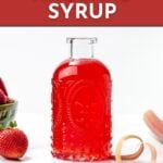 A glass bottle of bright red strawberry rhubarb syrup sits on a white surface, with fresh strawberries and rhubarb nearby. Text reads "Strawberry Rhubarb Syrup. Get the recipe at feastandwest.com".