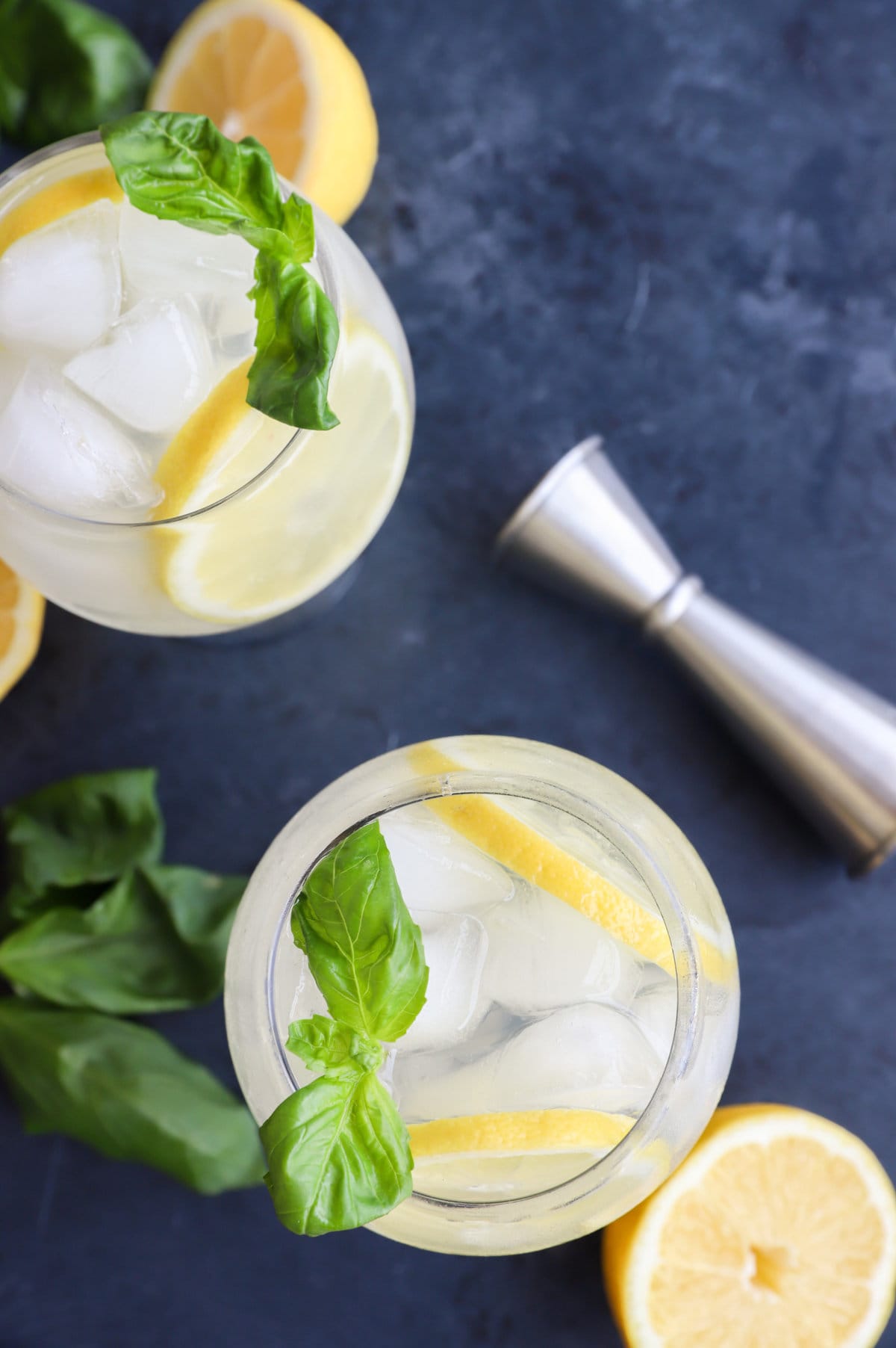 Two glasses of iced lemonade with lemon slices and basil leaves, surrounded by lemons and a metal jigger.