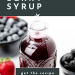 A bottle of Mixed Berry Syrup surrounded by fresh strawberries, blueberries, and blackberries on a white surface.