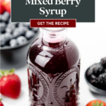 A bottle of Mixed Berry Syrup surrounded by fresh strawberries, blueberries, and blackberries.