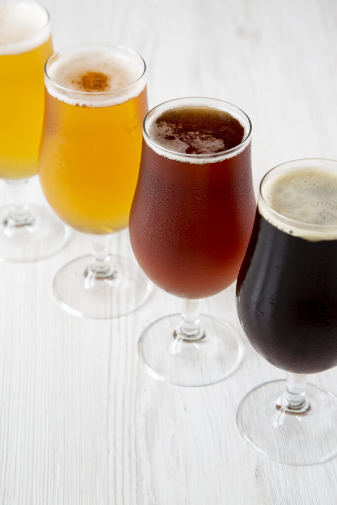 Four glasses of beer in varying shades from light yellow to dark brown, arranged in a diagonal line on a white surface.