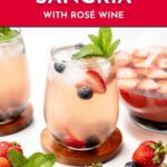 Glass of mixed berry sangria with ice, strawberries, blueberries, and mint, next to a pitcher and fresh berries.
