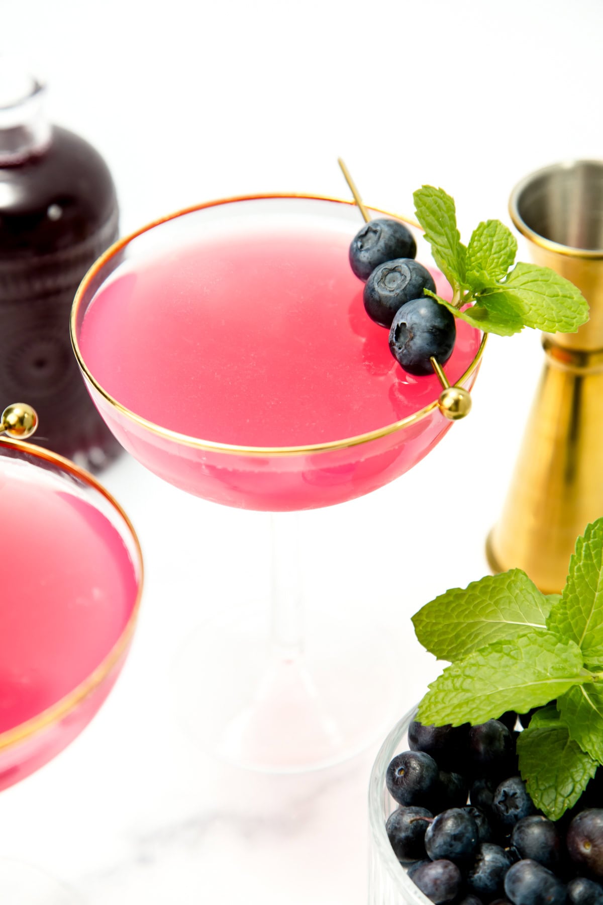 A pink cocktail in a coupe glass, garnished with blueberries and mint, with a gold jigger and fresh blueberries nearby.
