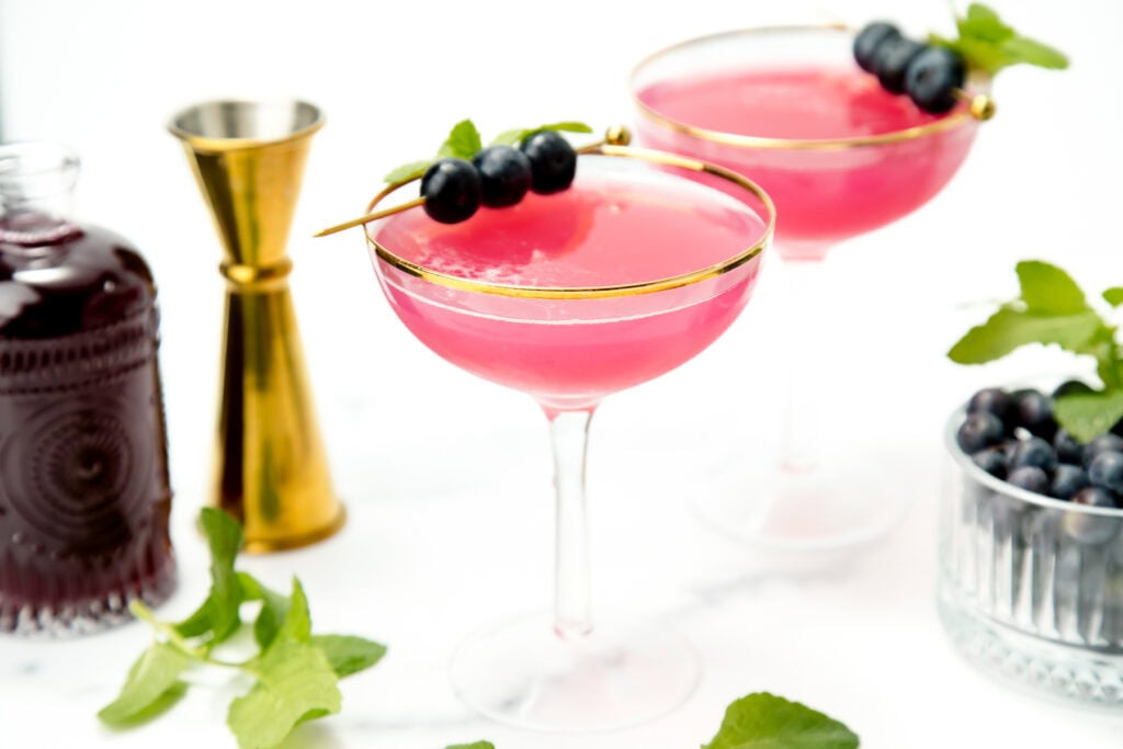 Two pink cocktails in coupe glasses, garnished with blueberries and mint, with bar tools and ingredients nearby.