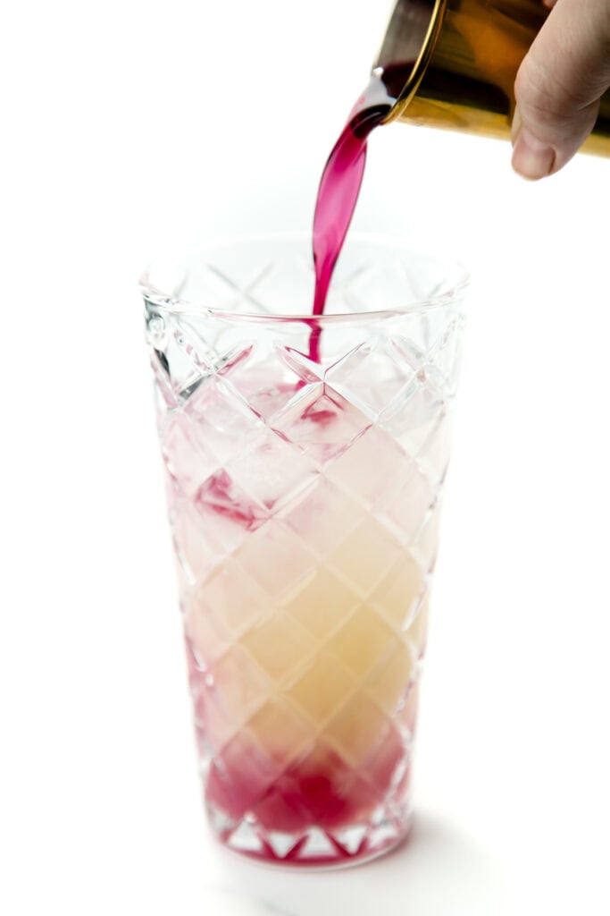 Red liquid being poured from a brass shaker into a tall glass of ice on a white background.