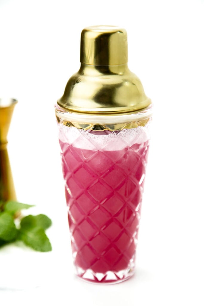 A glass cocktail shaker with a gold lid and pink drink inside, against a white background.