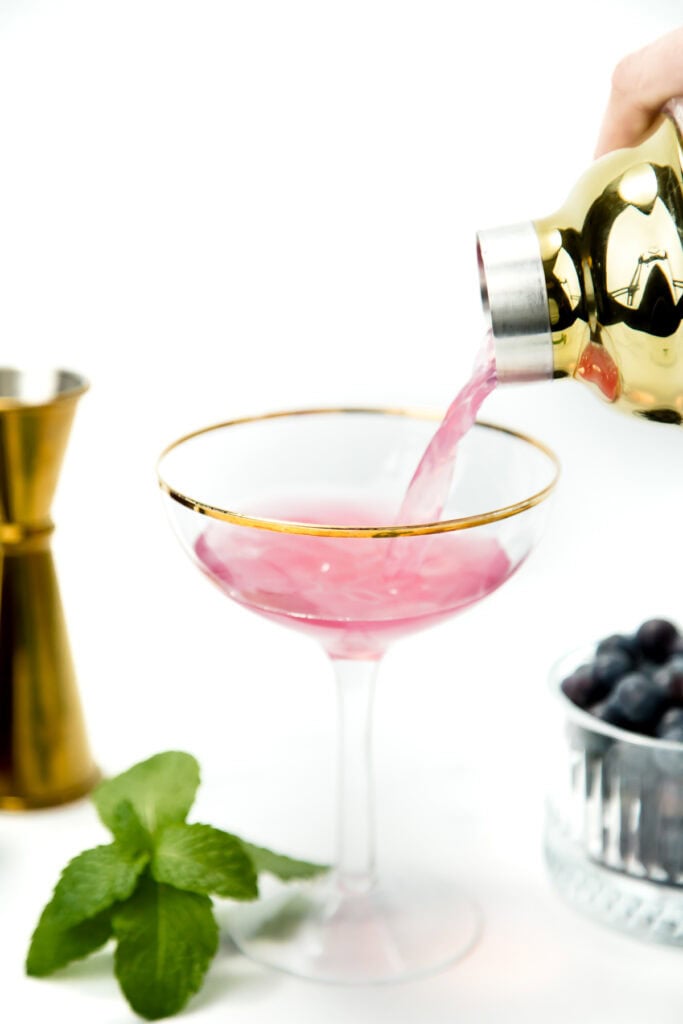 Pink cocktail being poured into a gold-rimmed glass, with mint and blueberries beside it on a white background.