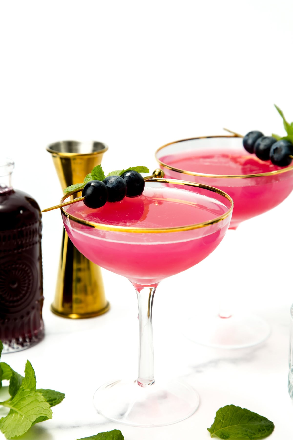 Two pink cocktails in coupe glasses, garnished with blueberries and mint, with a gold jigger and syrup bottle nearby.