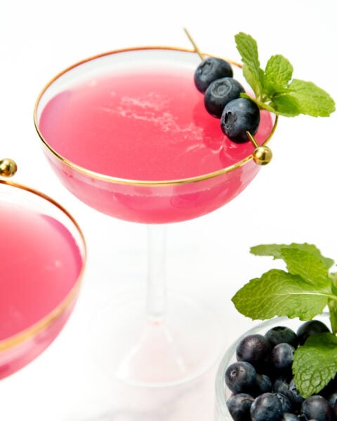 A pink cocktail in a gold-rimmed glass, garnished with blueberries and mint, beside a bowl of blueberries.