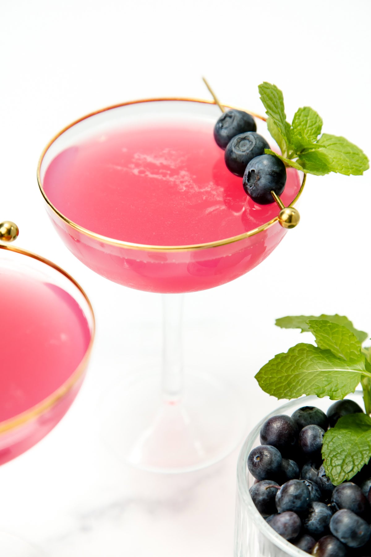 A pink cocktail in a gold-rimmed glass, garnished with blueberries and mint, beside a bowl of blueberries.