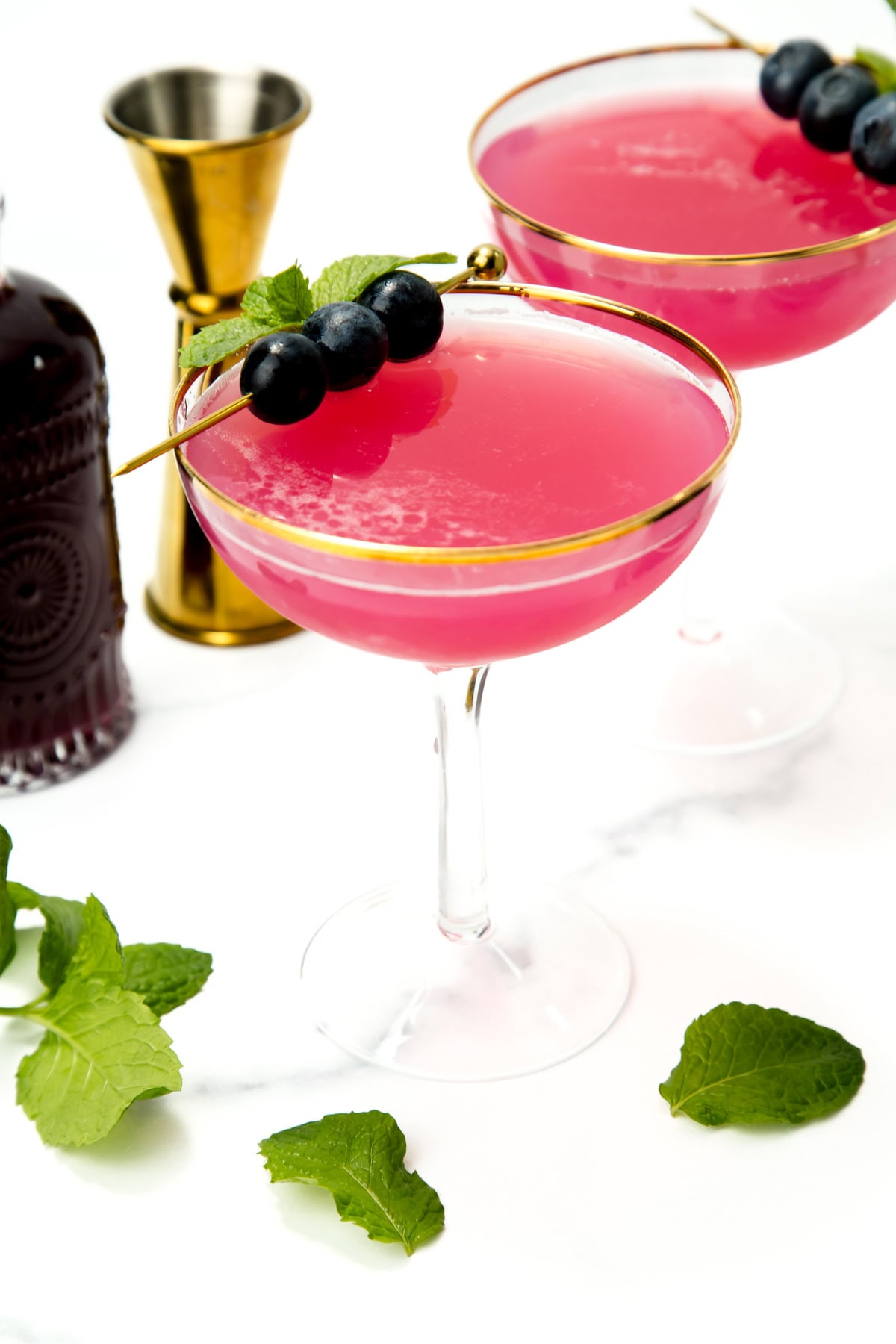 Two pink cocktails in gold-rimmed glasses, garnished with blueberries and mint, surrounded by mint leaves.