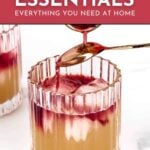 A spoon pours red syrup into a glass of cocktail; text reads "Home Bar Essentials: Everything You Need at Home. Learn More. feastandwest.com".